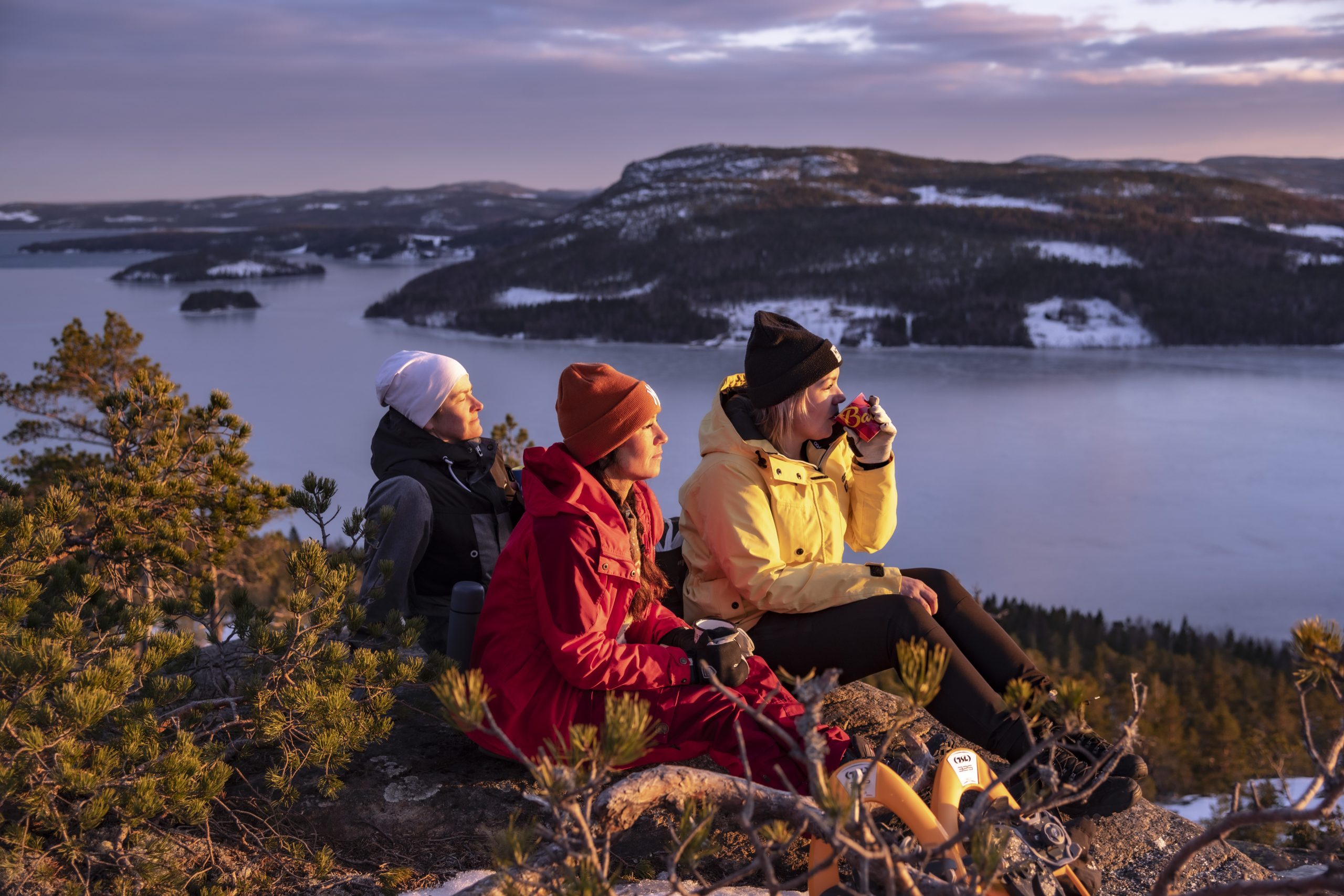 Tre vandrare fikar med utsikt över isbelagd fjärd och snöklädda berg vid solnedgång i Höga Kusten