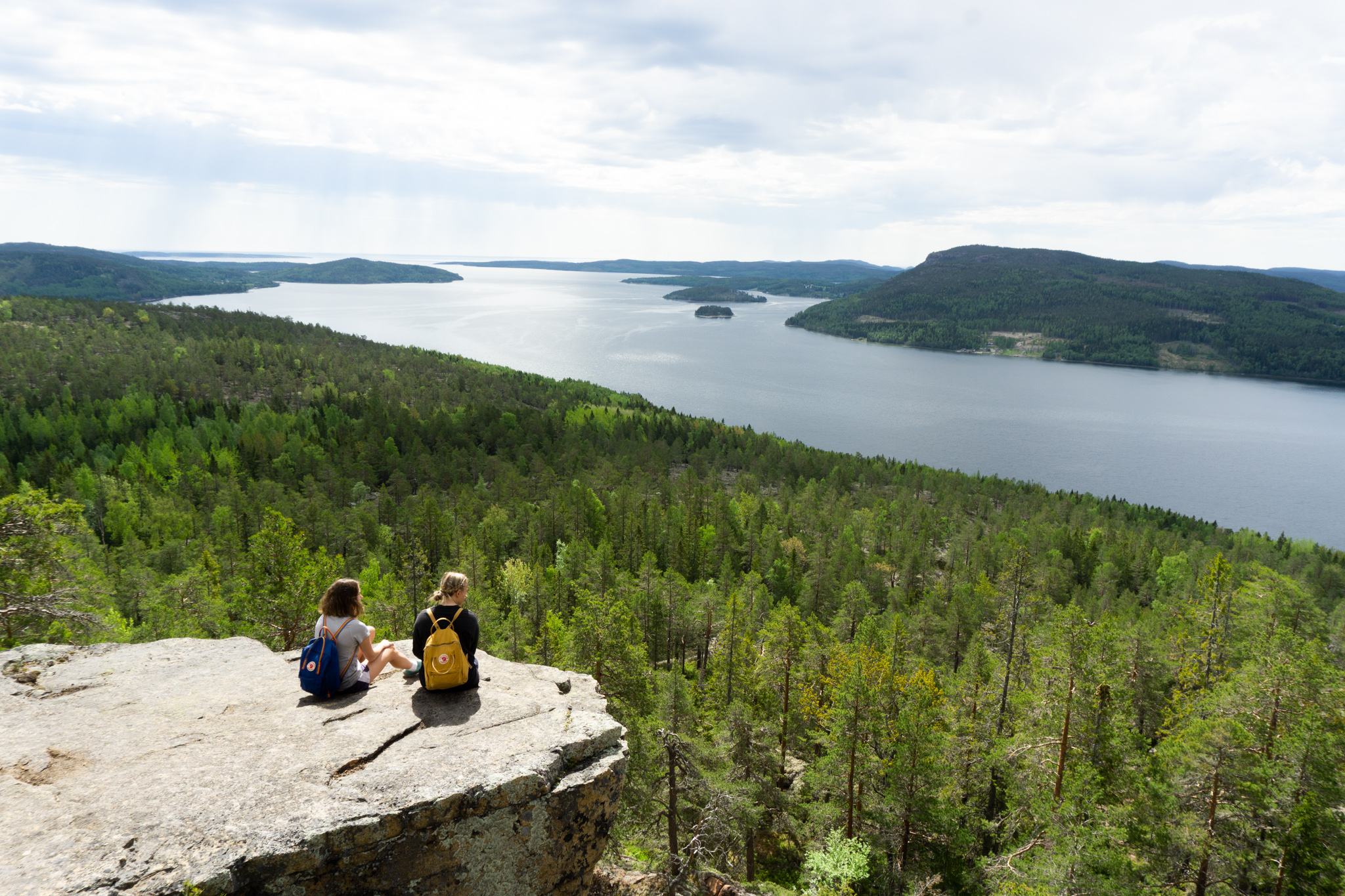 Två personer sitter vid Getsvedjebergets kant och blickar ut över skog, hav och öar i Höga Kusten