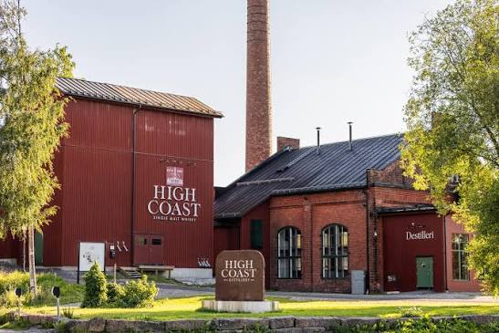 Exteriörvy av High Coast Whisky Distillery. Röda fabriksbyggnader, hög skorsten, gräsmatta och träd. Ådalsgården bra boende nära High Coast Whisky